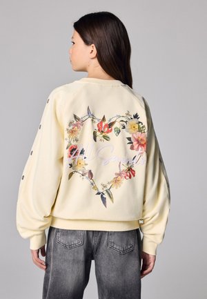 Mujer joven con cabello largo y castaño lleva una sudadera crema con un corazón floral y el texto "All Saints" en la espalda, combinada con vaqueros negros desgastados.