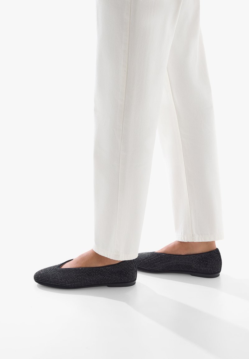 Pantofole nere testurizzate con punta affusolata, suola piatta e design superiore intrecciato abbinate a pantaloni bianchi.