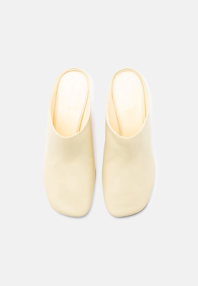 Κρεμ δερμάτινα σανδάλια χωρίς κορδόνια με τετράγωνες μύτες, επίπεδες σόλες και ελάχιστη λεπτομέρεια ραφής, απεικονιζόμενα από πάνω σε λευκό φόντο.