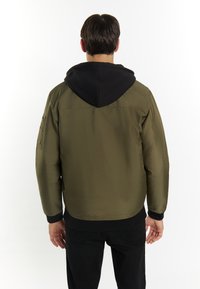 Giubbotto bomber verde oliva con felpa nera, polsini e fondo a coste, caratterizzato da una tasca laterale sulla manica sinistra. Design minimalista.
