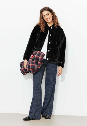 Femme souriante, portant une veste douce noire, un haut blanc, un pantalon large bleu, tenant une chemise à carreaux rouge et bleu contre un mur blanc.