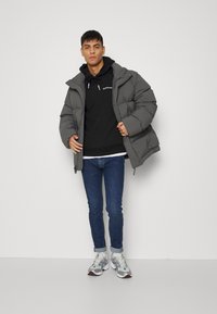 Casaco puffer cinzento com capuz, apresentando um fecho de correr e grandes bolsos, sobre um hoodie preto e combinado com jeans azuis justos e sapatilhas.