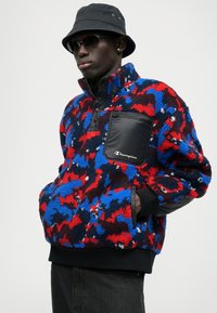 Fleece pullover in red, blue, and black pattern with a zippered chest pocket and a black nylon panel; features ribbed cuffs and hem.