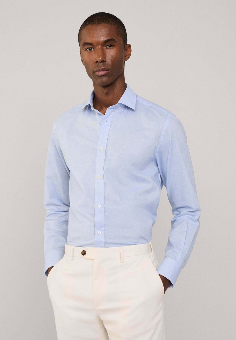Camisa azul claro abotonada en una tela suave y liviana con cuello puntiagudo, un bolsillo, y mangas largas, combinada con pantalones color crema.