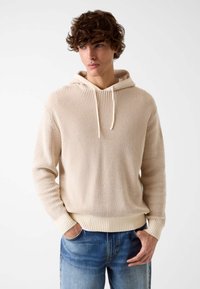 Felpa con cappuccio in maglia beige con polsini e orlo a coste, dotata di coulisse e superficie testurizzata. Abbinata a jeans chiari in denim.