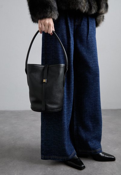 Saco de pele preto em forma de balde com detalhes em metal dourado, segurado por uma pessoa que está vestindo calças largas com padrão azul e sapatos pretos de bico fino.