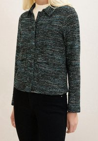 Femme portant un cardigan boutonné en noir et bleu texturé par-dessus un haut blanc, associé à un pantalon noir, se tenant contre un fond neutre.