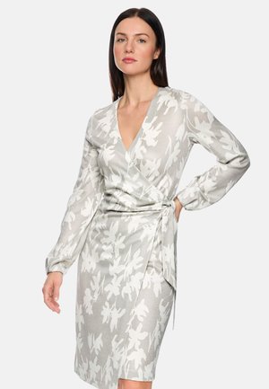 Woman wearing a long-sleeve, V-neck wrap dress with white floral patterns on a light gray background, standing with one hand on hip.