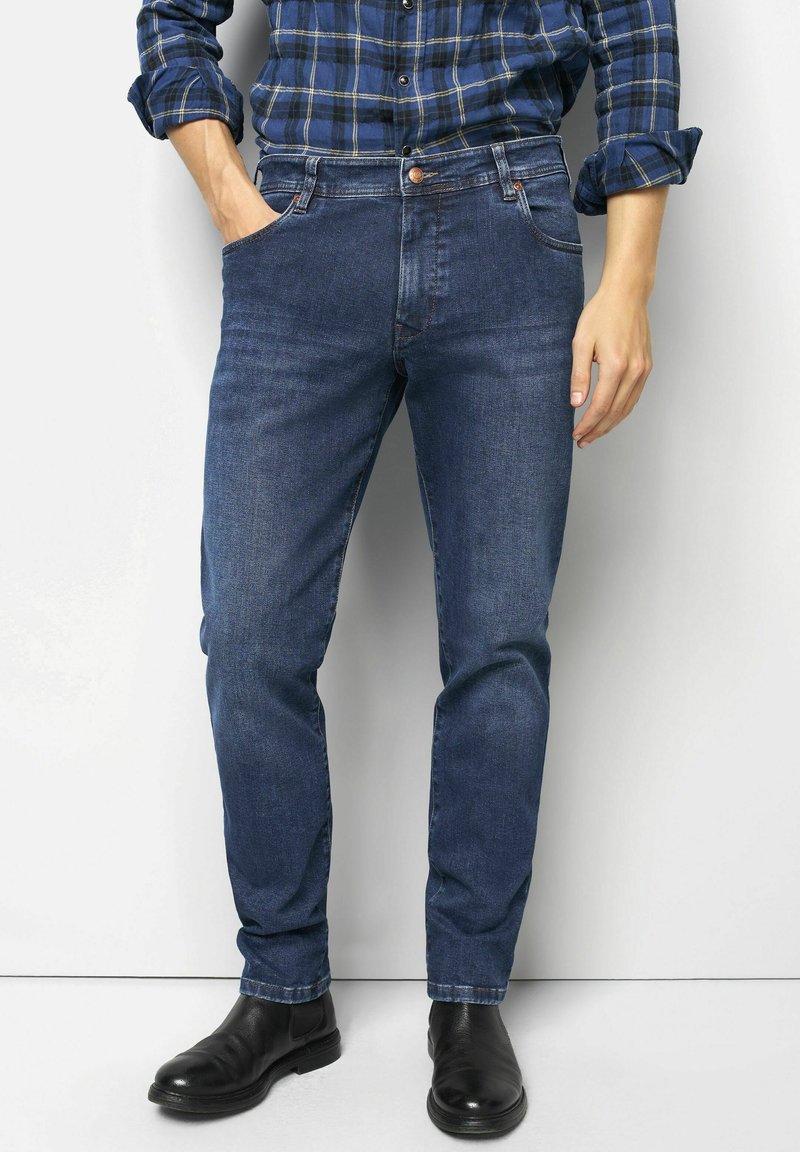 Donkerblauwe spijkerbroek met een recht model. Kenmerkt zich door zakken, een knoopsluiting en subtiele stikseldetails. Gecombineerd met een geruite blouse en zwarte laarzen.