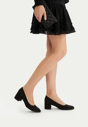 Žena nosící černé boty na podpatku s blokem a černou krajkovou sukni, držící černé prošívané psaníčko.