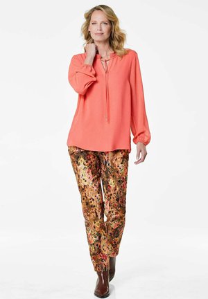 Blouse corail avec de longues manches bouffantes et un détail à nouer, associée à un pantalon aux motifs dorés et floraux. Bottes à talons hautes marron.