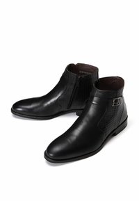 Botas de tobillo de cuero negro con un panel texturizado, cierre de cremallera y una correa decorativa, con un diseño elegante y punta afilada.