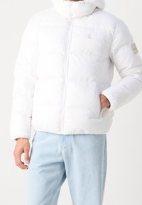 Biała kurtka puchowa z kapturem i kieszeniami na zamek, noszona z jasnoniebieskimi dżinsami, ręka w kieszeni, z logo "CK" na klatce piersiowej i rękawie.