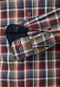 Koszula w kratę z mieszanką czerwonych, niebieskich i zielonych kwadratów. Ciemnoniebieskie mankiety z detalami w postaci guzików oraz kontrastującą podszewką.