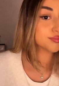 Gros plan sur une personne avec des cheveux bruns ondulés de longueur moyenne. Elle porte un pull blanc et un délicat collier en or avec un pendentif en forme de goutte.