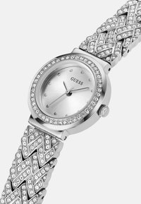 Sølv ur med en glansfuld rund urskive, udsmykket med klare krystaller omkring bezellet og et dekorativt, vævet rem.