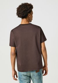 Maglietta in cotone marrone con collo a giro, maniche corte e vestibilità rilassata, abbinata a jeans in denim azzurri chiari con una toppa.