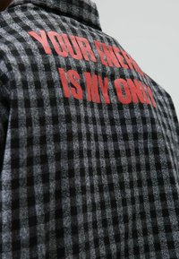 Grijs-zwarte geruite flanellen blouse met rode tekst op de achterkant die zegt "JOUW ENERGIE IS MIJN ENIGE." Voorzien van een overhemdkraag.