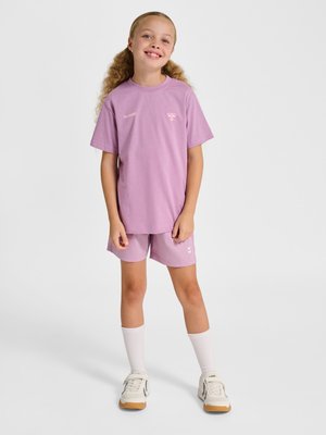 Jong meisje met krullend haar, die een lila T-shirt en korte broek, witte sokken en witte sneakers draagt, staat tegen een effen achtergrond.