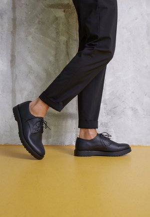Osoba nosząca czarne eleganckie buty i czarne spodnie z mankietami, podnosi jedną nogę stojąc na żółtej podłodze przy szarej ścianie.