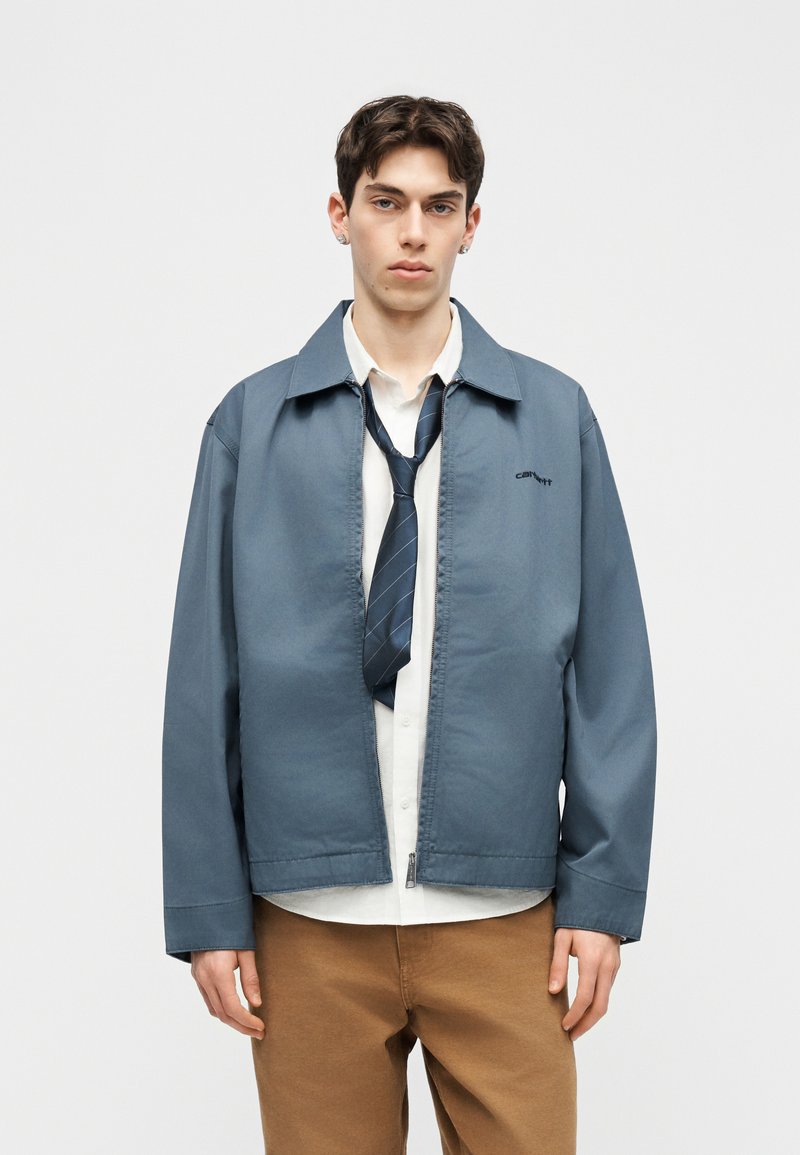 Giovane uomo con capelli corti e scuri che indossa una giacca blu sopra una camicia bianca e una cravatta blu navy allentata, abbinata a pantaloni marroni, su uno sfondo semplice.