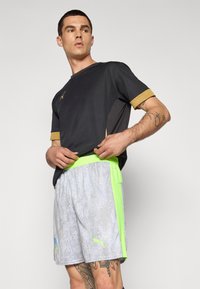 Sort træningsskjorte med guld accenter, parret med lysegrå shorts med en grøn talje og vandsymbol mønster.