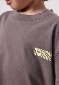 Bambino che indossa una maglietta marrone con scritta gialla "AMERICAN VINTAGE" sul petto, che mostra il collo e parte del viso con una luce soffusa.