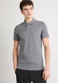 Sivý polo tričko vyrobené z bavlny, s klasickým golierom, dvojitou gombíkovou légou a malou nášivkou s logom na ľavej hrudi.