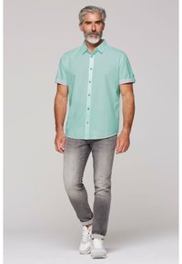 Camicia button-up verde menta a maniche corte con i polsini arrotolati, abbinata a jeans grigi sbiaditi e sneakers bianche. Design semplice e look casual.