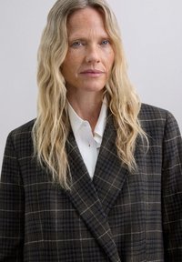 Blazer marrón de cuadros con un corte holgado, adornado con acentos blancos y un cuello en V profundo, usado sobre una camisa blanca de botones. Fondo difuminado.