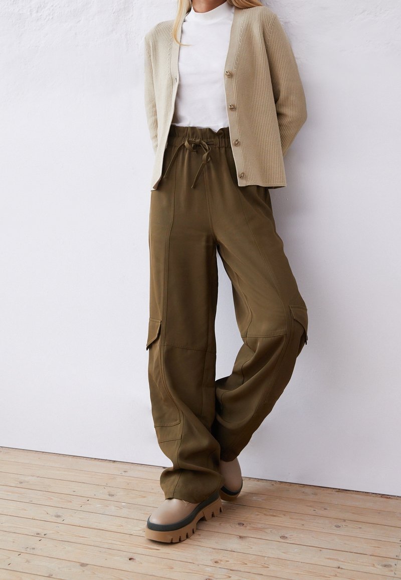 Femme portant un cardigan beige, un col roulé blanc, un pantalon cargo kaki à jambes larges et des chaussures beiges à semelle épaisse, debout contre un mur blanc.
