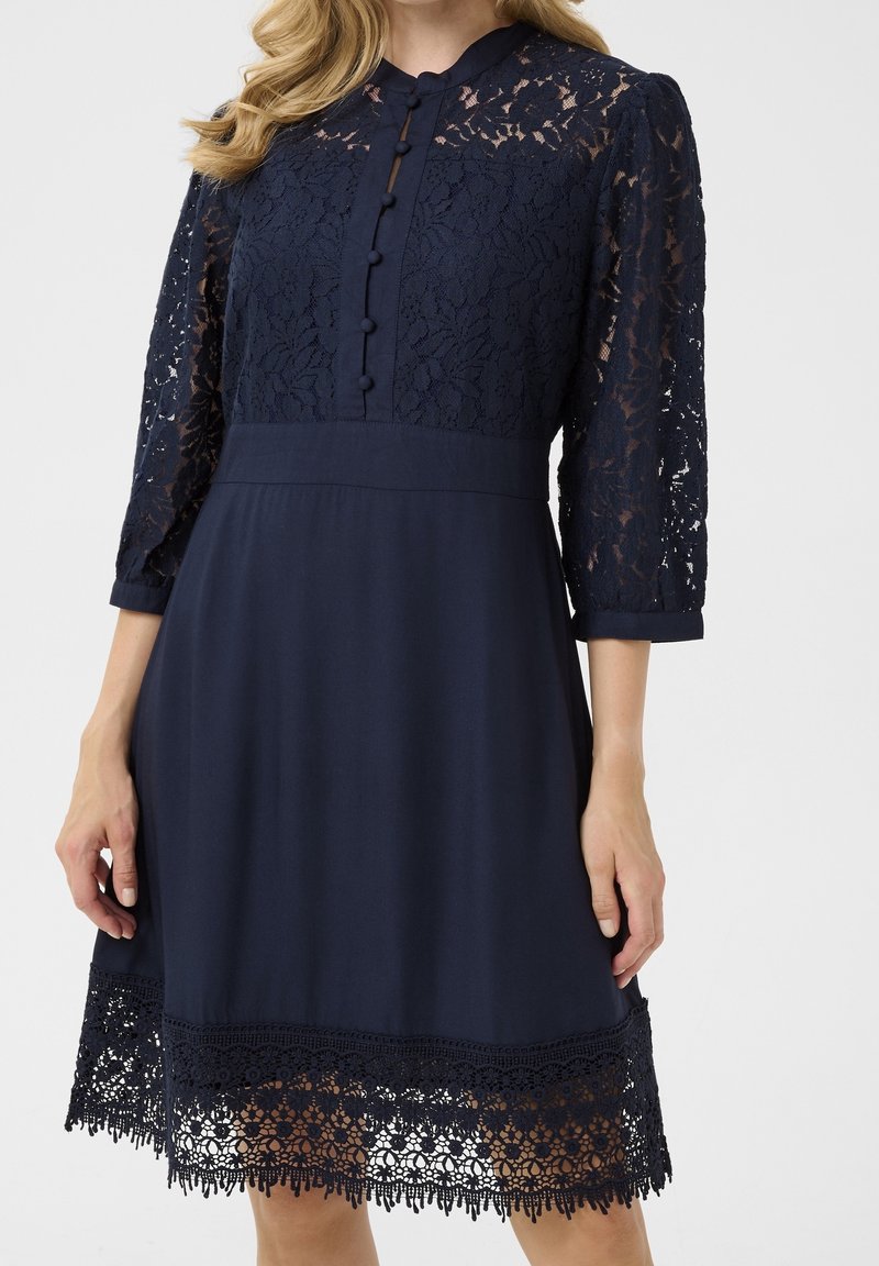 Robe bleu marine avec corsage en dentelle florale, manches trois-quarts en dentelle, boutons recouverts de tissu et bordure en dentelle au niveau de l'ourlet jusqu'au genou.