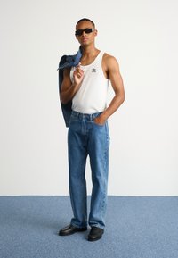 Bela rebrasta majica bez rukava z logotipom v črni barvi, svetlo modri kavbojki, jeans jakna čez ramo, črne slip-on čevlje, stoji na modri teksturirani tleh.