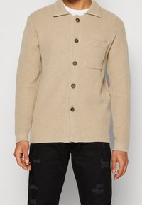 Hombre vistiendo una camisa de botones de manga larga texturada en color beige con bolsillo en el pecho y pantalones negros desgastados, de pie contra un fondo liso.