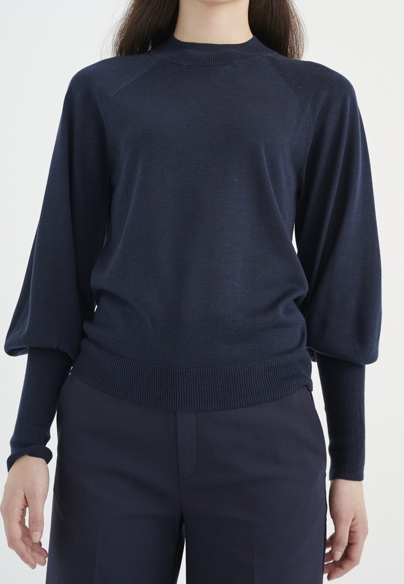 Granatowy dzianinowy sweter z wysokim kołnierzem, rękawami raglanowymi oraz ściągaczami na mankietach i dole. Miękka tekstura o luźnym kroju.