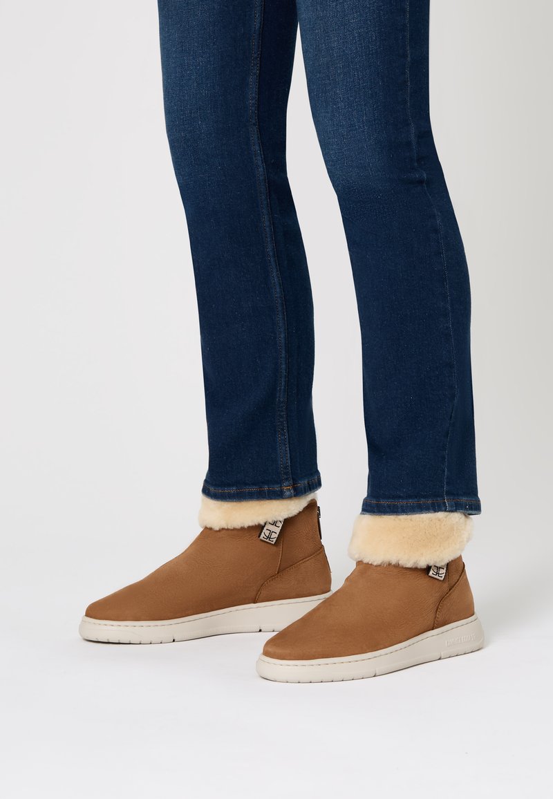 Stivaletti in camoscio beige con foderatura in pelo crema, suola in gomma bianca e zip laterale, indossati con jeans blu scuro.