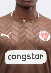 Κοντινή λήψη καφέ ποδοσφαιρικής φανέλας FC St. Pauli με γεωμετρικό μοτίβο, λογότυπο Puma, σήμα συλλόγου και κείμενο χορηγού "congstar" στο μπροστινό μέρος.