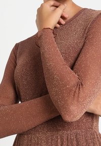 Hnědý lesklý top s dlouhým rukávem, přiléhavého střihu, texturované látky a nařasenými manžetami. Jemný lesk od zlatých nití.