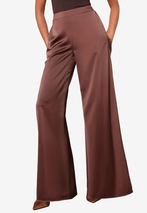 Mujer con pantalones de satén marrón de tiro alto y pierna ancha con bolsillos laterales, combinados con zapatos marrones de punta puntiaguda sobre un fondo liso.