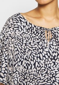 Blouse à motifs noir et blanc avec un col rond, ornée d'un petit trou en forme de clé relié par des cordons en tissu assortis.