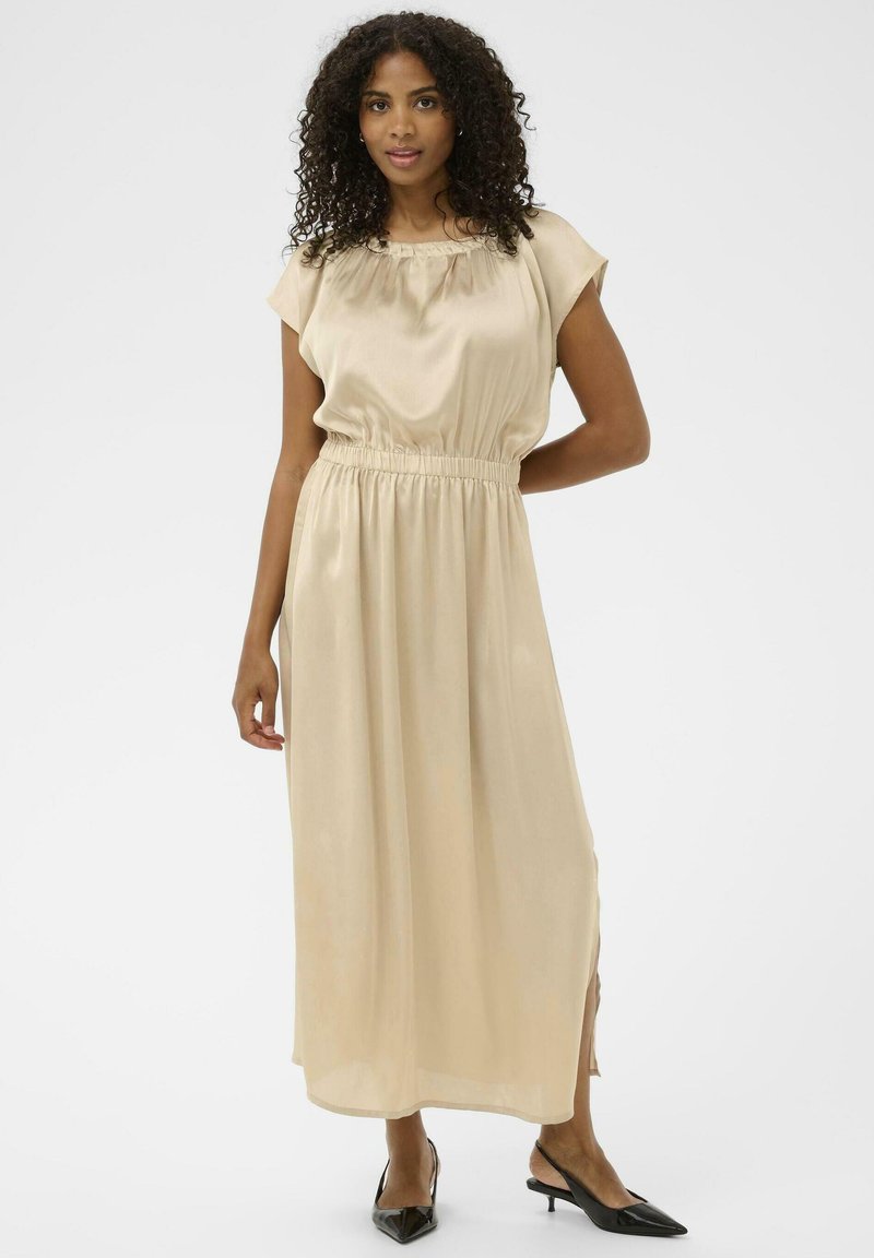 Abito lungo in satin beige con maniche corte, fascione elastico in vita e spacchi laterali, abbinato a décolleté nere a punta. Tessuto liscio.
