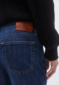 Pantalones vaqueros de mezclilla en azul oscuro con superficie texturizada, con un parche de cuero Calvin Klein en la parte trasera y costuras contrastantes en los bolsillos.