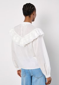 Weiße Bluse mit einem großen, bestickten Rüschensaum und langen Ärmeln, kombiniert mit hellblauen Jeans.