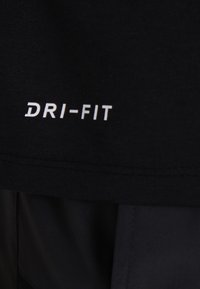 Černá látka DRI-FIT, hladká textura, atletický střih, s bílým potištěným logem na spodním lemu. Měkký a lehký materiál.