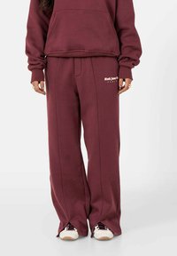 Persoon in maroon wijde sweatpants met de tekst "Sixth June Co Paris" op de linkerdij en witte sneakers met maroon veters.
