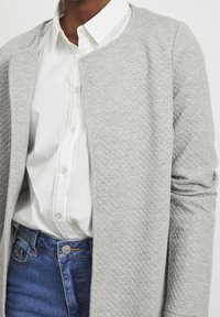 Cardigan gris texturé avec un col rond, porté sur une chemise blanche à boutons. Taille en denim visible avec détail de bouton.