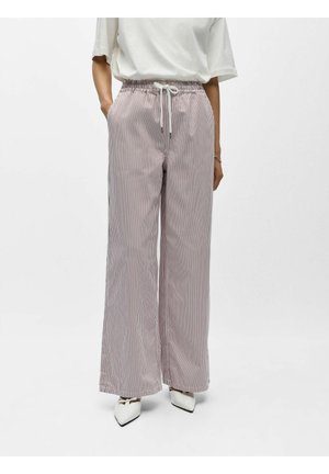 Person trägt weit geschnittene, hoch taillierte gestreifte Hosen mit Kordelzug, weißen Oberteil und spitze weiße Schuhe, steht vor einfachem Hintergrund.