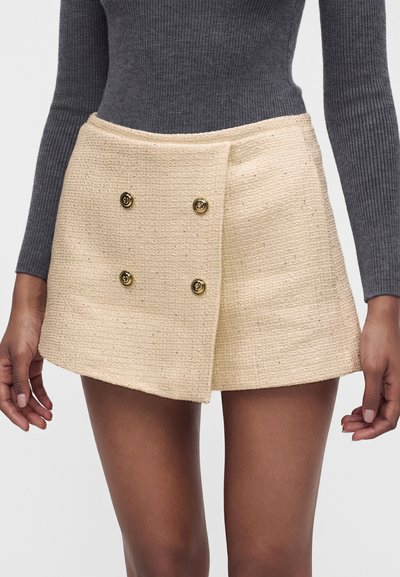 Gros plan d'une personne portant une mini-jupe beige texturée avec quatre boutons dorés et un haut à manches longues côtelé gris foncé.