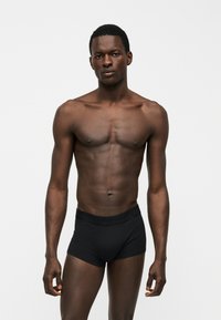 Αθλητικός άνδρας με σκουρόχρωμο δέρμα, κοιτάζει μπροστά, φοράει μαύρα εφαρμοστά μποξεράκια μπροστά σε απλό ανοιχτό γκρι φόντο.