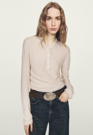 Top rosa chiaro a maniche lunghe con scollo henley e dettagli con bottoni, abbinato a jeans scuri e una cintura marrone con fibbia ovale argentata.